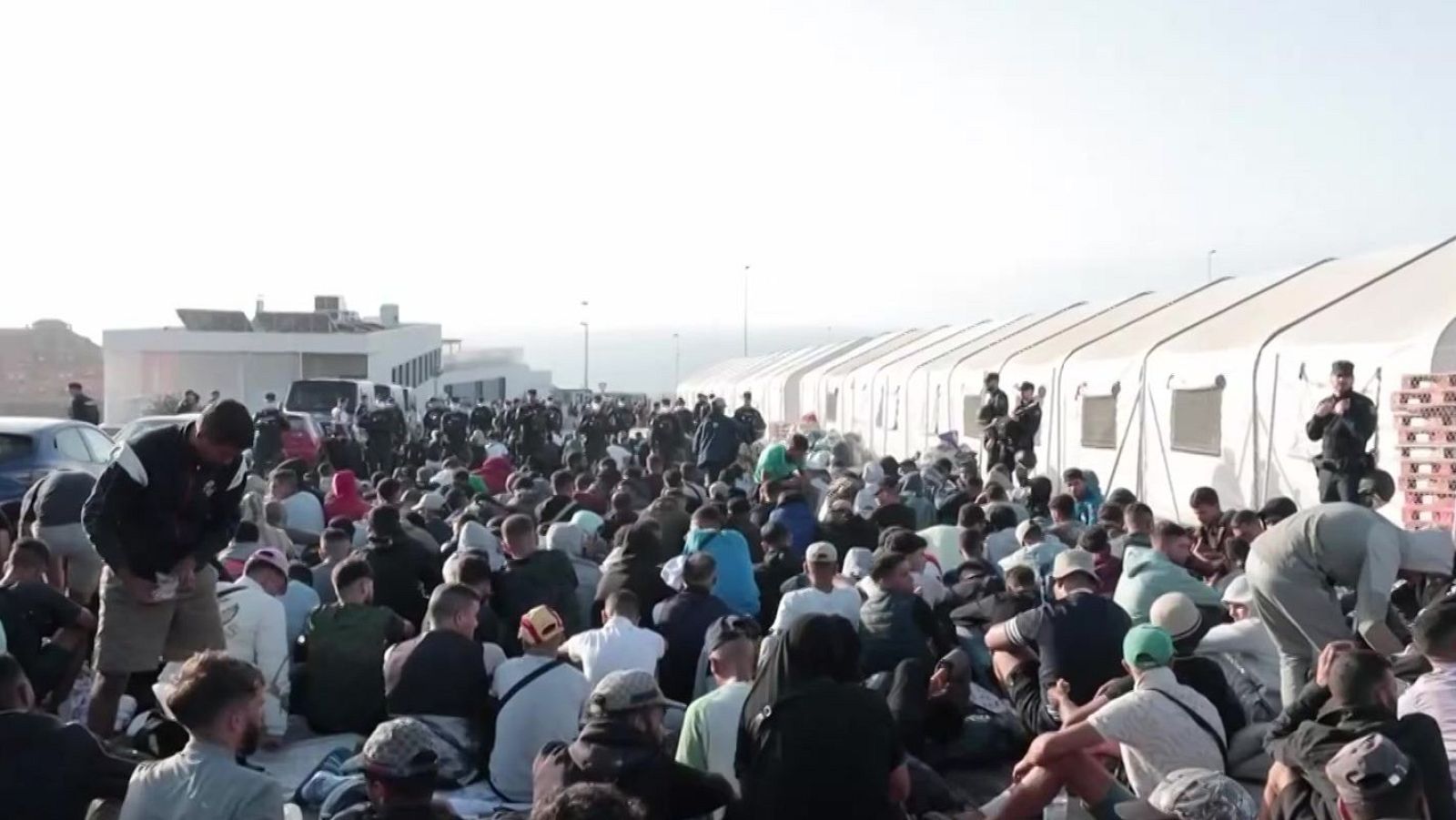 Miles de personas, predominantemente hombres jóvenes, sentados en un campamento al aire libre bajo el sol. Se observan tiendas blancas, personal uniformado, edificios y vehículos en el fondo.
