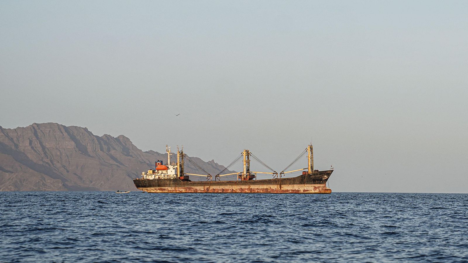 Gran barco comercial con casco oscuro y óxido, superestructura blanca, grúas amarillas y botes naranjas, fondeado en mar azul con montañas áridas al fondo.