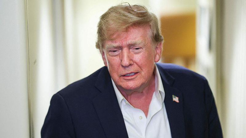 Donald Trump, con traje azul marino y camisa blanca, mira hacia abajo con expresión seria. Luce un pin de la bandera de EE.UU. en su solapa. Fondo interior desenfocado.
