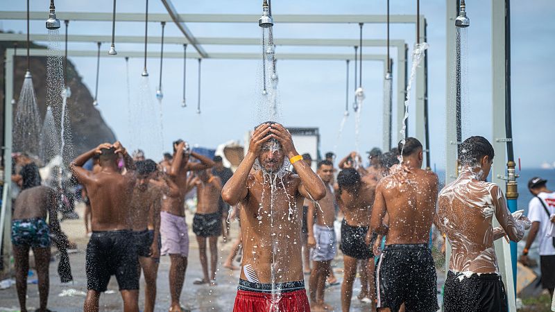 Hombres jóvenes se duchan al aire libre en una estructura temporal. Uno de ellos, en el centro, viste bañador rojo y tiene un peine visible. Al fondo, una colina rocosa y el mar.