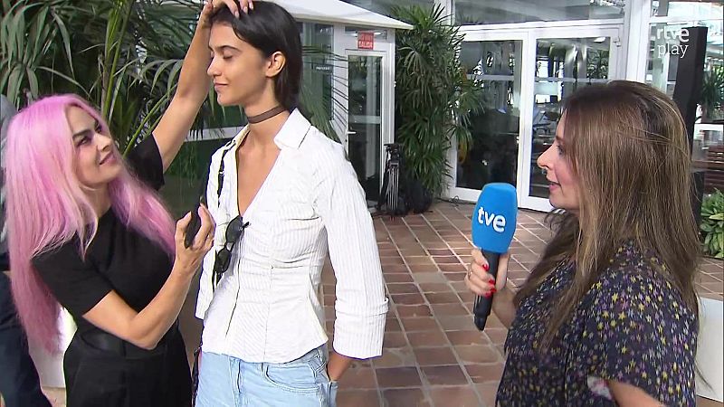 Estilista de cabello rosa ajusta a modelo con gafas de sol y choker, mientras reportera de TVE con micrófono se acerca. Fondo con plantas y señal de "SALIDA".