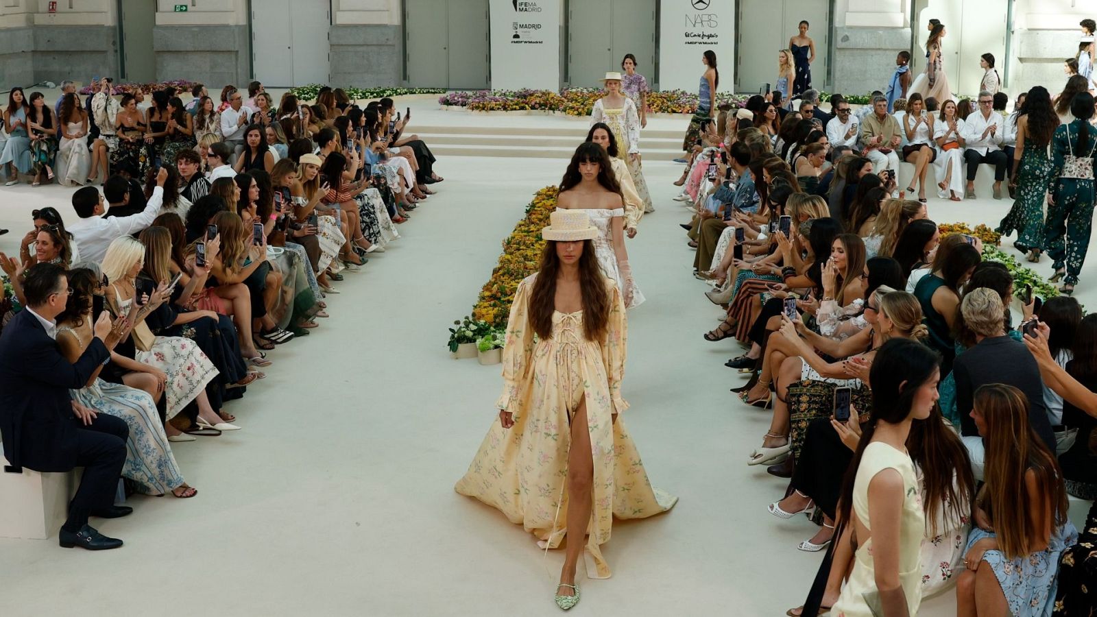 Desfile de moda en la Mercedes-Benz Fashion Week Madrid con modelo en conjunto amarillo pálido y sombrero, público observando y grabando.