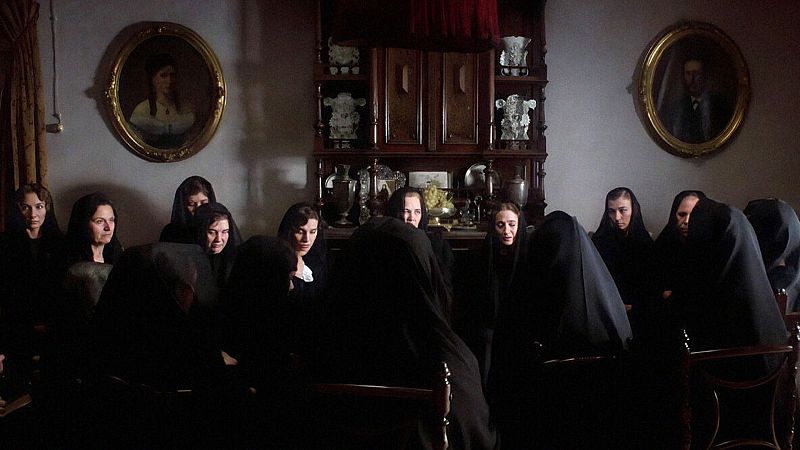 Diez mujeres de luto riguroso, incluyendo a Ana Belén, Enriqueta Carballeira, Vicky Peña y Aurora Pastor, se reúnen en un salón austero con retratos familiares y un gran aparador.
