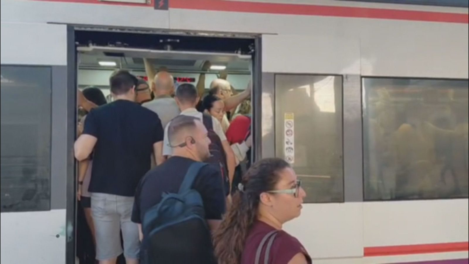 Tren de cercanías blanco con franja roja, puerta abierta y vagón repleto de gente. Mujer con gafas de sol y hombre con mochila azul en la entrada. Alta afluencia de pasajeros.