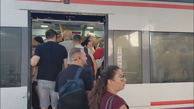 Tren de cercan�as blanco con franja roja, puerta abierta y vag�n repleto de gente. Mujer con gafas de sol y hombre con mochila azul en la entrada. Alta afluencia de pasajeros.