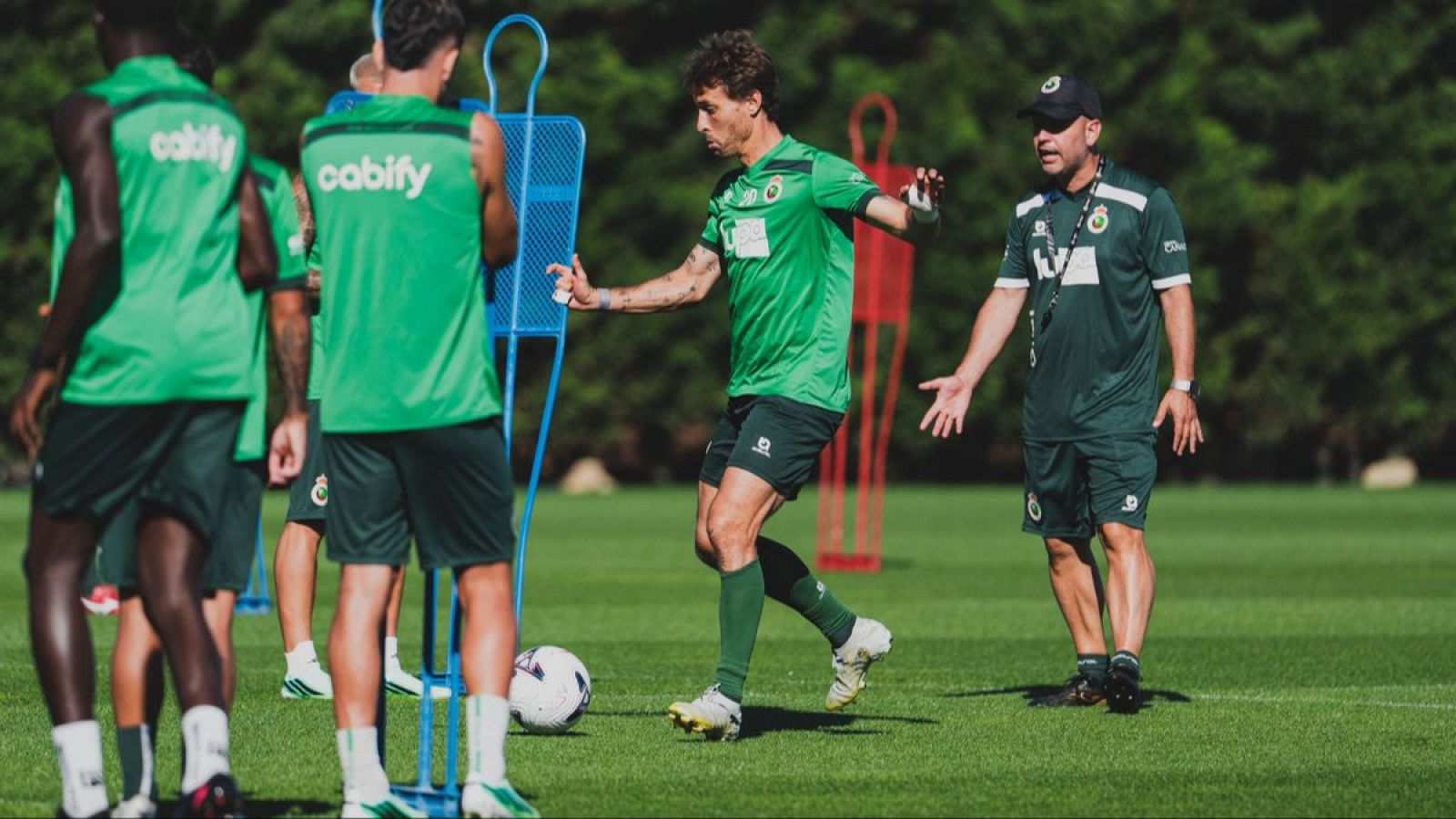 Íñigo Vicente practica control de balón en césped natural, supervisado por el entrenador José Alberto López. Chalecos con "cabify" y maniquíes de entrenamiento visibles.