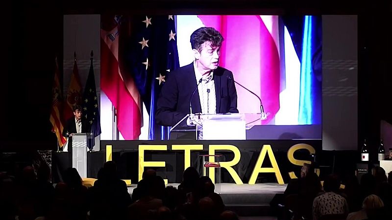 Santiago Auserón recibe el Premio de las Letras Aragonesas en un evento formal. Su imagen se proyecta en pantalla junto a la bandera de la UE, mientras un orador habla en vivo con las banderas de Aragón, España y la UE detrás.