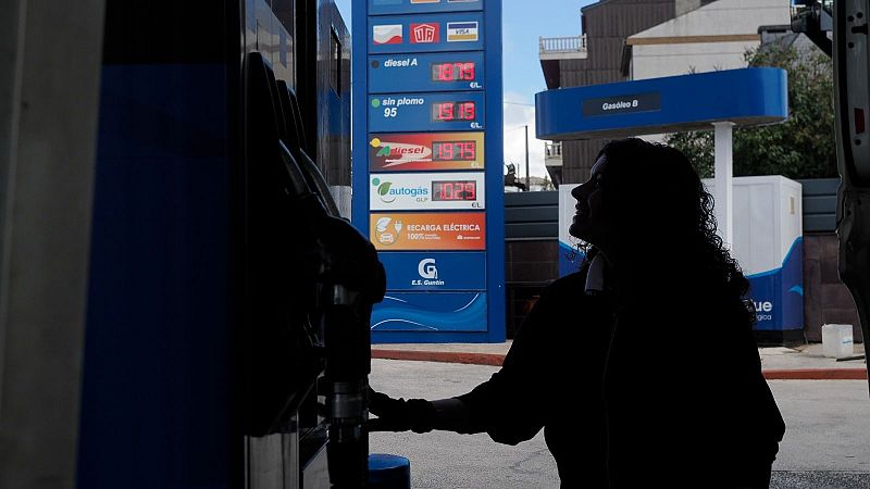 Persona en silueta en gasolinera, mirando precios de combustible. Poste azul muestra 'diesel A' 1.679€, 'sin plomo 95' 1.716€, 'diesel e+' 1.799€, 'autogas GLP' 0.929€ y 'RECARGA ELÉCTRICA 100%'.
