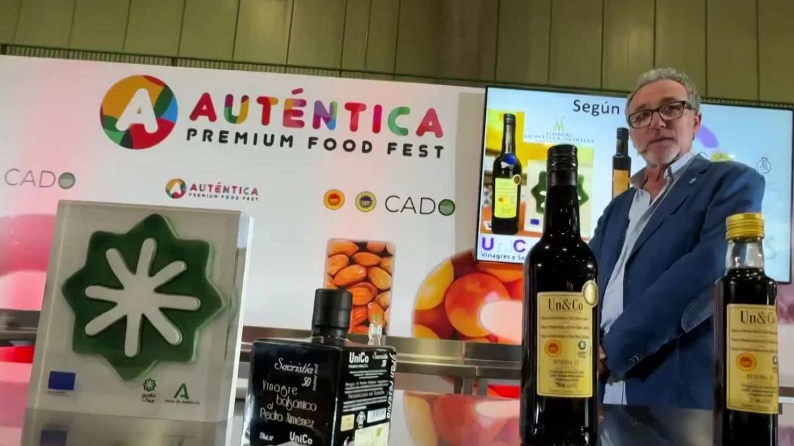Juanma Moreno, Presidente de la Junta de Andalucía, en el "Auténtica Premium Food Fest" junto a productos "Un&Co" y distintivos de la UE y la Junta.