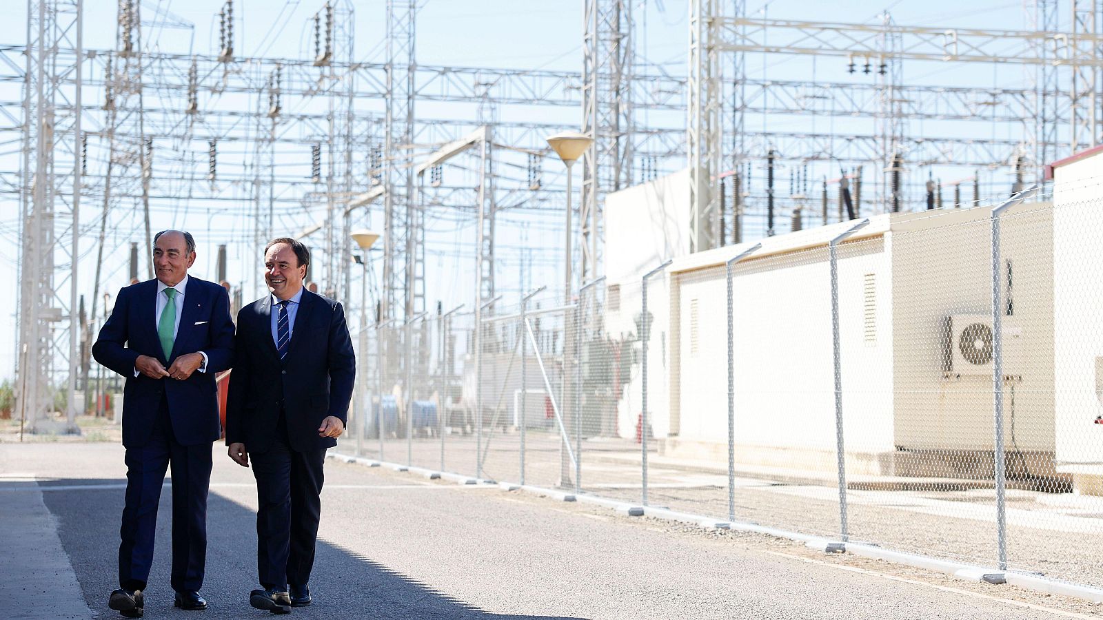 José Manuel Entrecanales Domecq y Alberto Núñez Feijóo, vestidos de traje, caminan por un camino asfaltado frente a una subestación eléctrica bajo un cielo despejado.