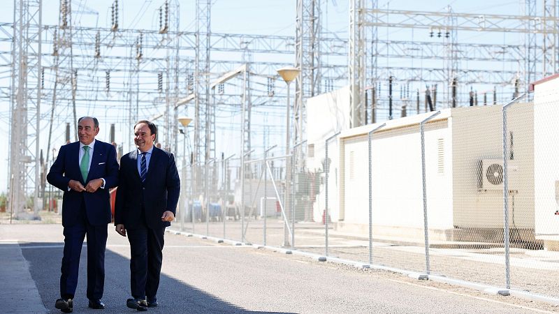 José Manuel Entrecanales Domecq y Alberto Núñez Feijóo, vestidos de traje, caminan por un camino asfaltado frente a una subestación eléctrica bajo un cielo despejado.