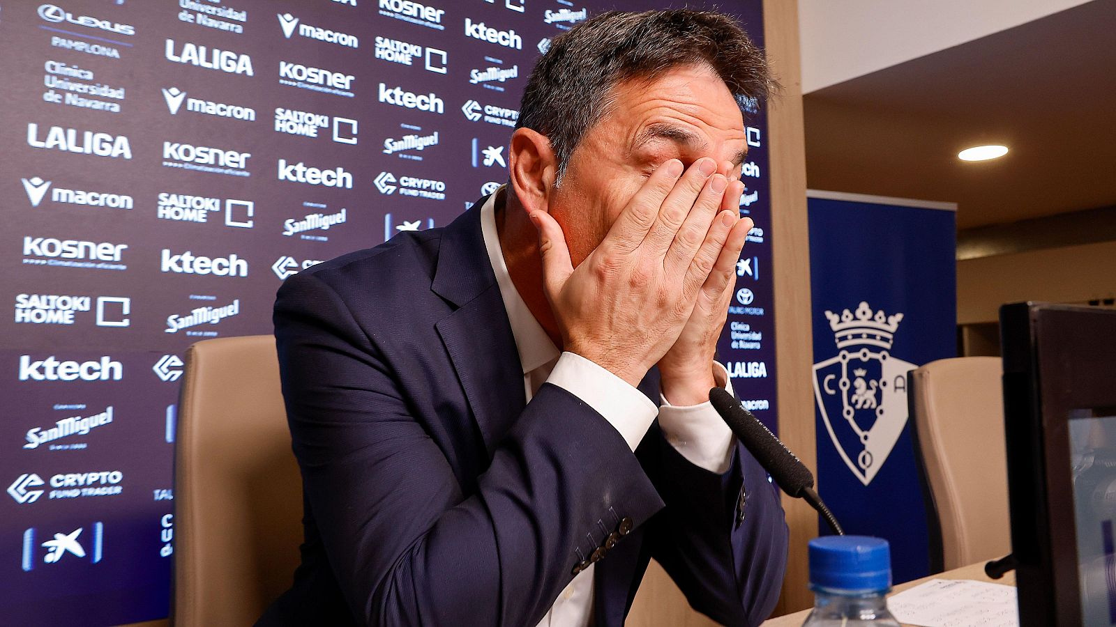 Primer plano de Braulio Vázquez, director deportivo del Osasuna, con las manos en el rostro en un gesto de emoción, rodeado de patrocinadores y el escudo del club.