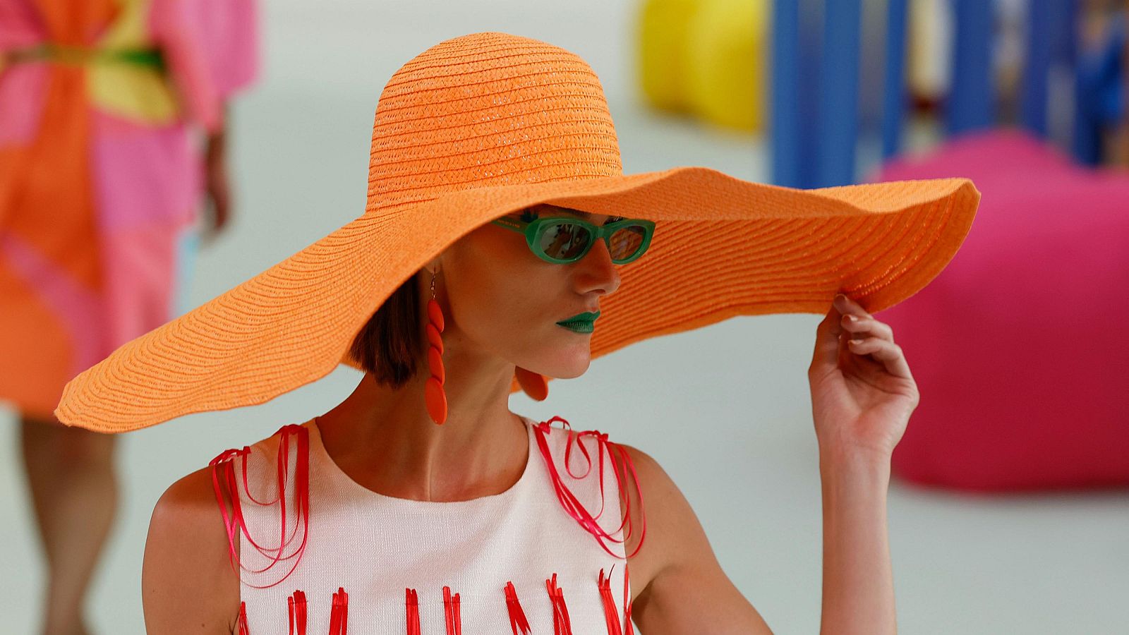 Modelo con sombrero naranja, gafas verdes y top blanco con cintas rojas, posa en un evento de moda con fondo colorido y abstracto.