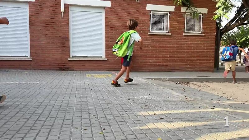 Niños corriendo sobre adoquines grises frente a un edificio de ladrillo. Uno lleva una mochila verde, el otro una con Sonic. Logo de '1' en la esquina inferior derecha.