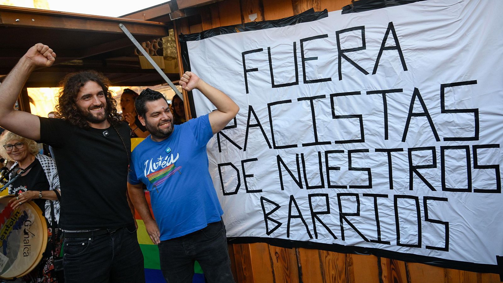 Pablo Fernández Santos y Serigne Mbayé Róson, con puños en alto, sonríen ante una pancarta antirracista. Una mujer con tambor visible a la izquierda.