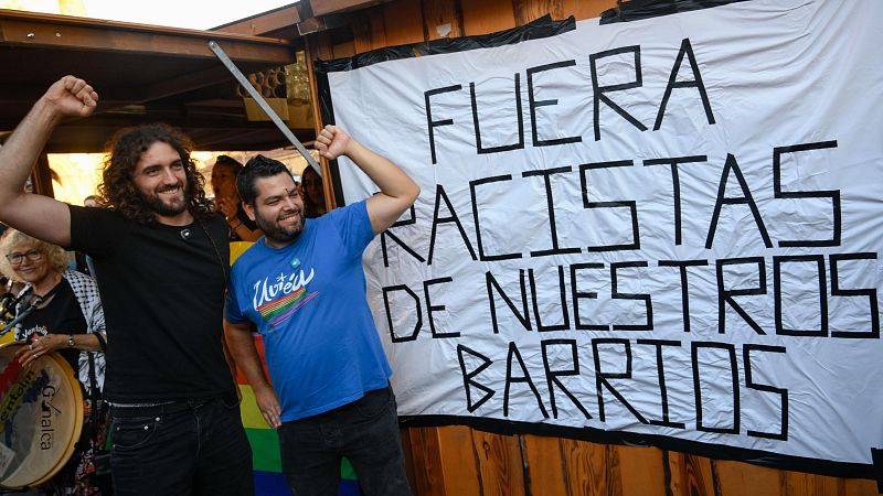 Pablo Fernández Santos y Serigne Mbayé Róson, con puños en alto, sonríen ante una pancarta antirracista. Una mujer con tambor visible a la izquierda.