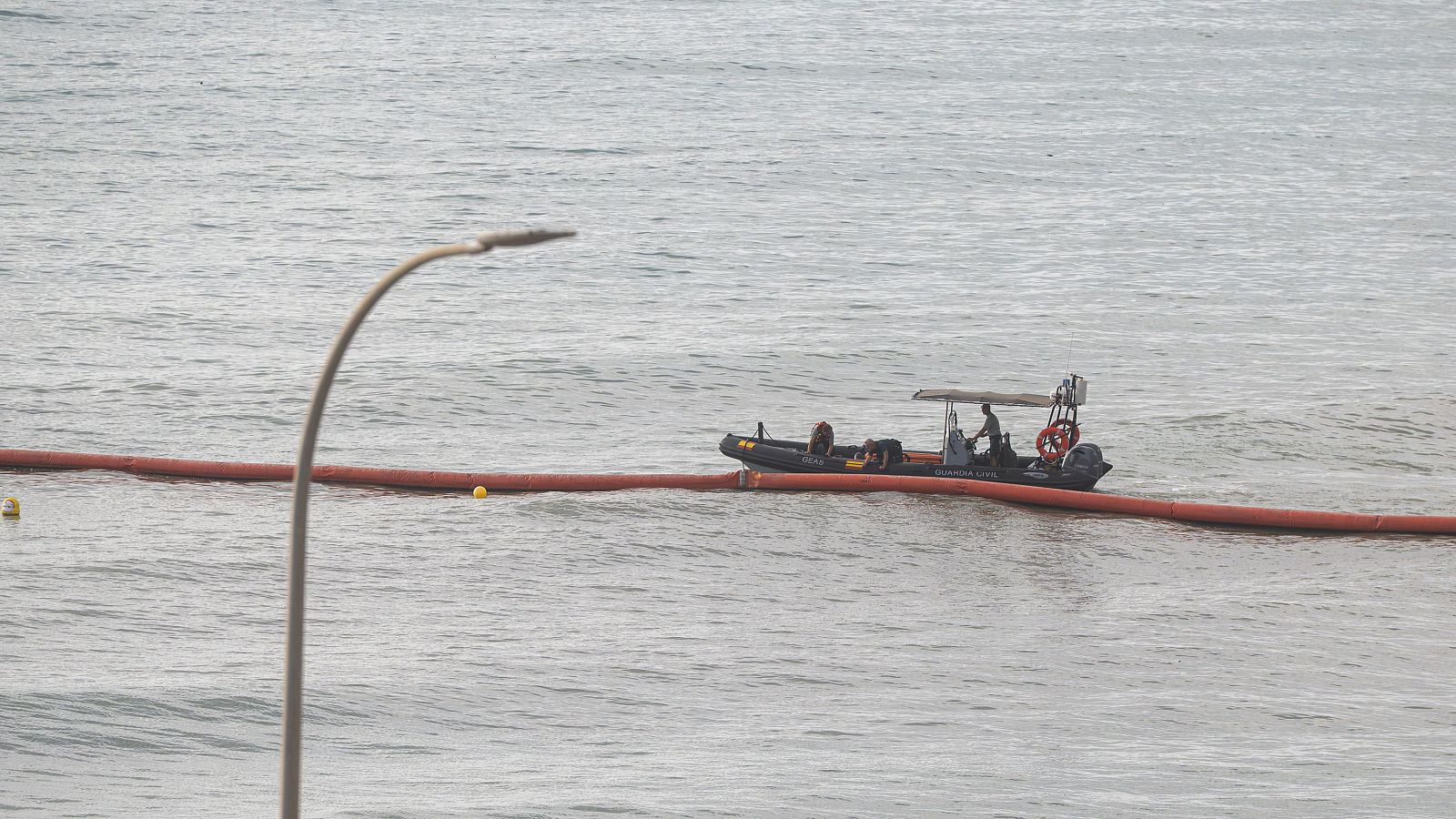 Lancha motora oscura con toldo y personas navega en mar gris. Barrera flotante naranja con boyas amarillas. Poste de alumbrado público curvado en primer plano.