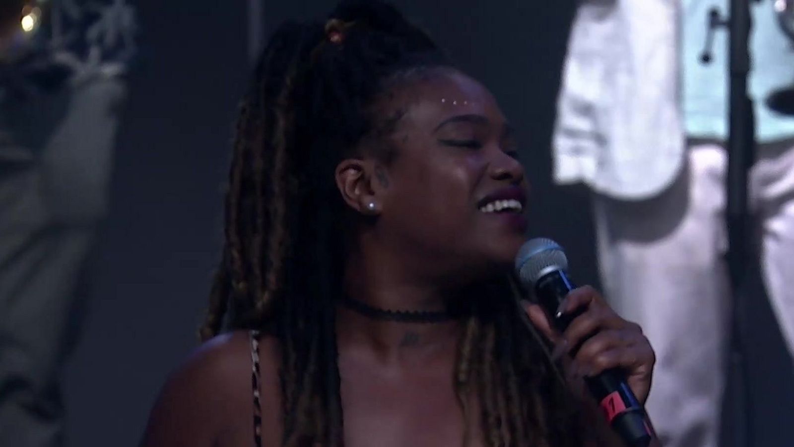 Cantante de piel oscura con dreadlocks y adornos brillantes en la sien, actuando en vivo con un micrófono negro. Expresión de concentración y emoción.