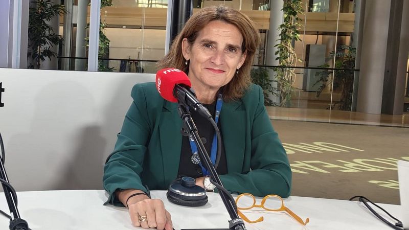 Primer plano de Teresa Ribera sonriendo a cámara, con un micrófono de RNE y gafas naranjas sobre la mesa. Fondo moderno con vegetación.