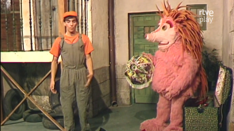 Espinete, personaje de pelaje rosa, sostiene una piñata colorida junto a un joven con mono gris y gorra naranja. Fondo de estudio con logo de RTVE Play.