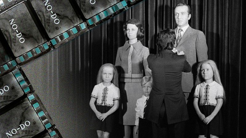 Tira de película del NO-DO sobre una escena de maniquíes familiares. Una persona real ajusta el traje del maniquí masculino. Iluminación uniforme sobre figuras rígidas.