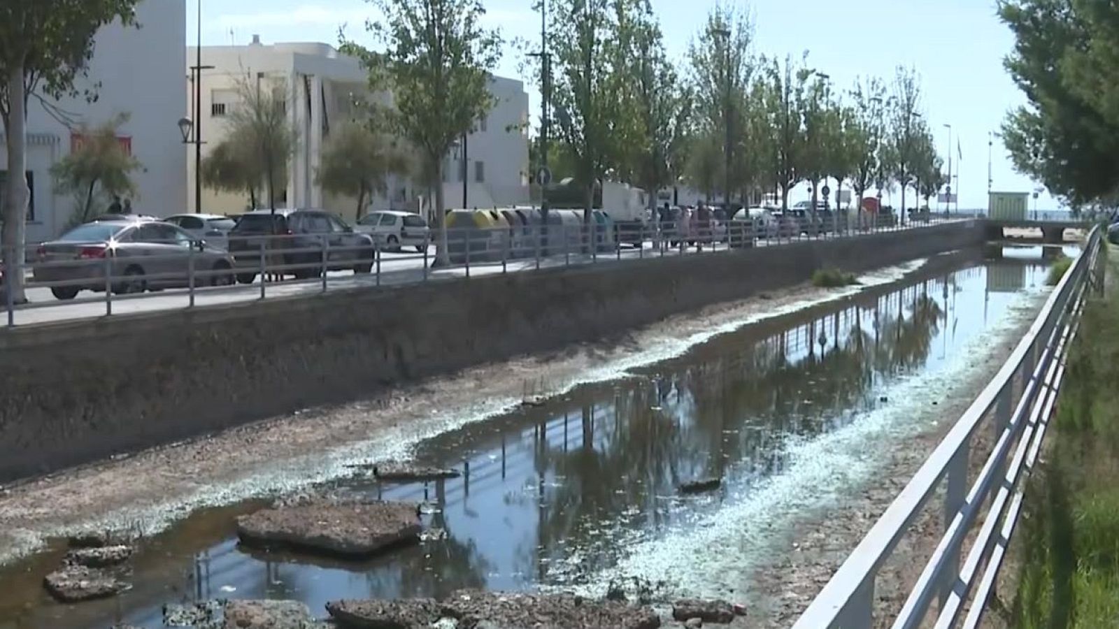 Canal con agua baja y vegetación, flanqueado por carretera con coches, contenedores de reciclaje, edificios y árboles. Un puente cruza el canal al fondo.