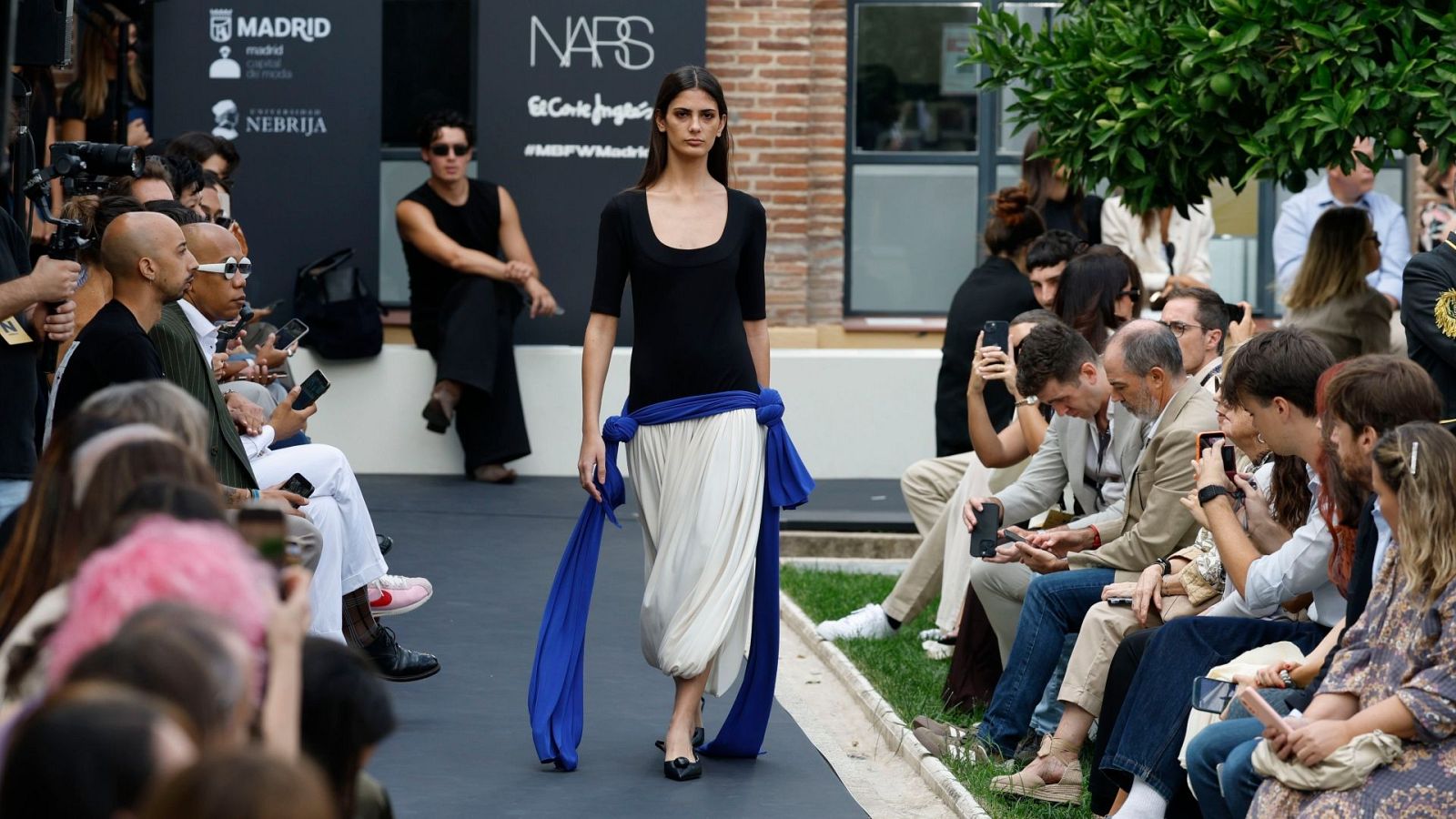 Modelo con camiseta negra y pantalón blanco roto, bufanda azul eléctrico. Audiencia con Pelayo Díaz y Omar Montes. Logotipos de Madrid, Nebrija, NARS, El Corte Inglés y #MBFWMadrid al fondo.