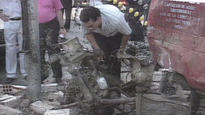 Hombre con gafas inspecciona restos de vehículo entre escombros. Pieza roja con texto: "CAPTACION DE AGUAS SUBTERRANEAS", "AVD DE LA LIBERTAD 75-2", "TLF: 965 / 78705", "CREDA".