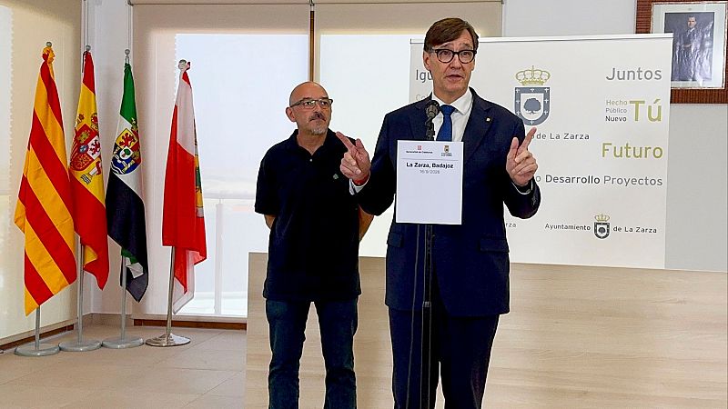 Salvador Illa habla en La Zarza, Badajoz, el 16-9-2024, con banderas y un banner del Ayuntamiento de La Zarza de fondo. Un hombre con polo azul le acompaña.