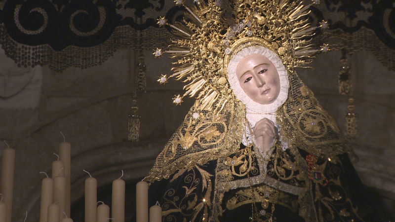 Escultura de la Virgen Dolorosa con manto y saya oscuros bordados en oro, corona dorada con rayos y estrellas, manos juntas en oración, velas de cera y fondo oscuro.