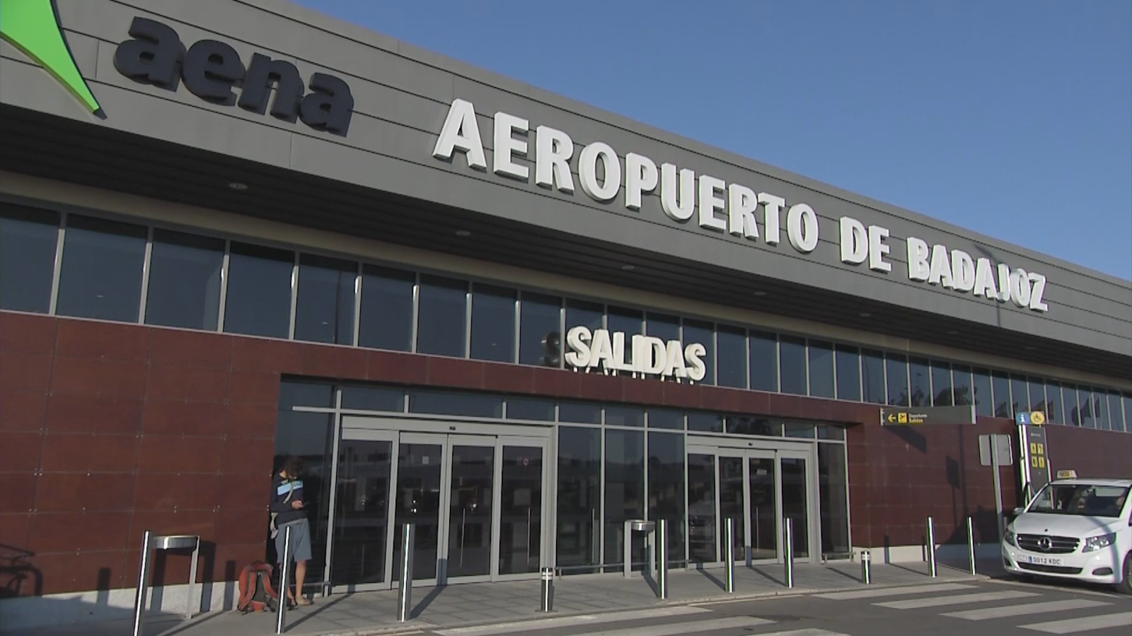 Fachada del Aeropuerto de Badajoz con "SALIDAS" visible. Una persona con mochila naranja y una furgoneta Mercedes-Benz blanca están presentes.