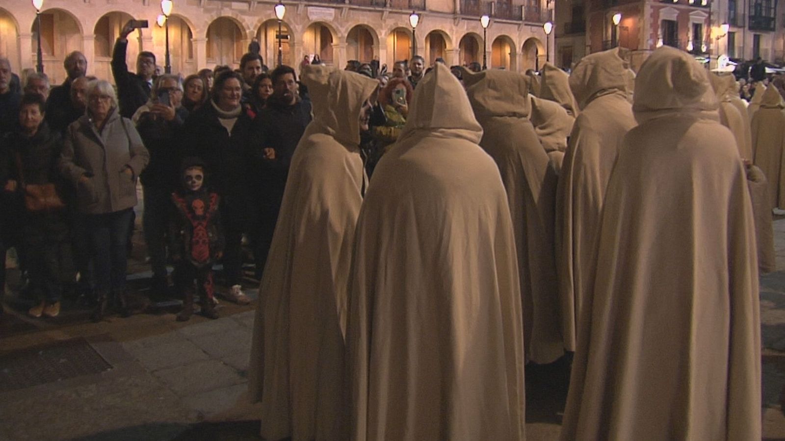 Procesión nocturna en Soria con figuras encapuchadas y espectadores, incluyendo un niño disfrazado de esqueleto, ante edificios históricos iluminados.