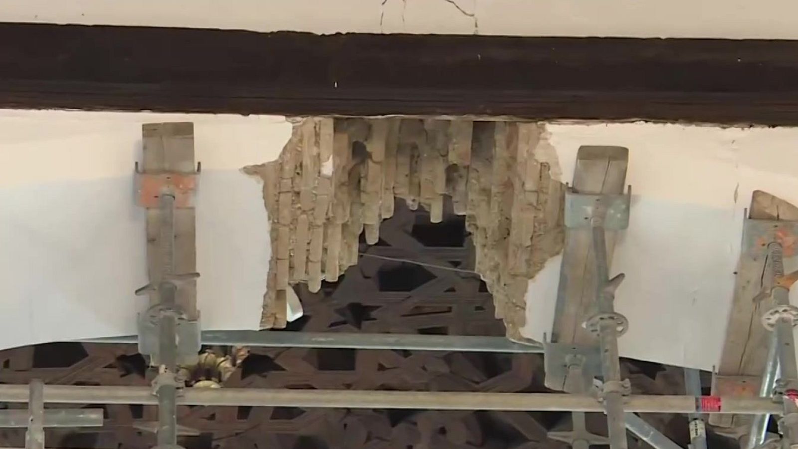 Techo interior dañado con viga de madera, superficie enlucida agrietada, listones de madera expuestos y andamio metálico debajo.