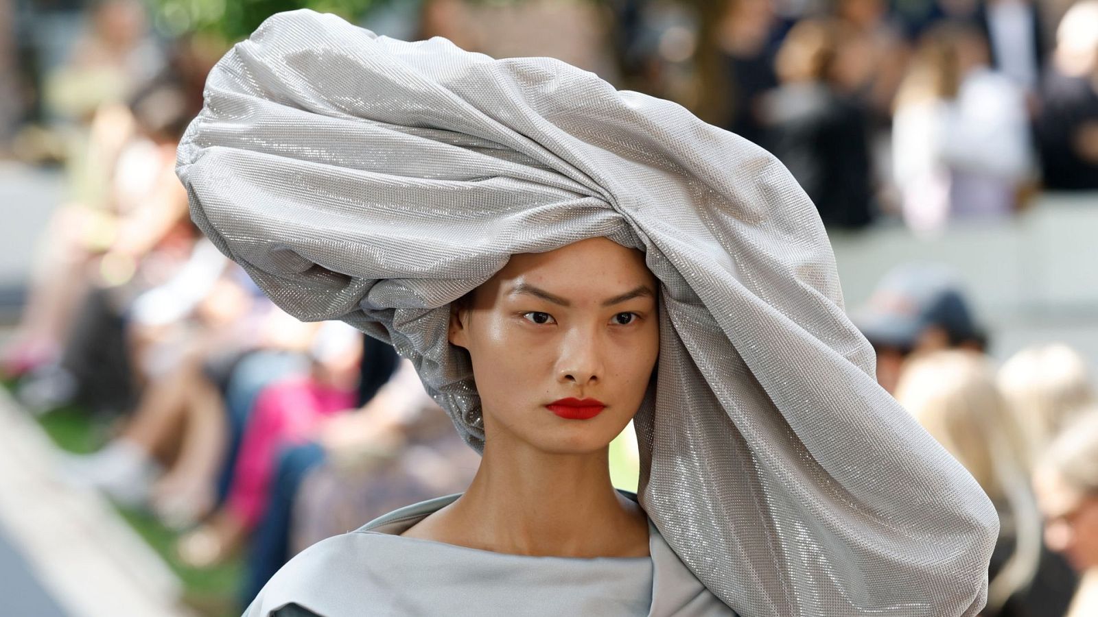 Modelo con tocado escultural gris plateado y labios rojos intensos en un evento de moda.