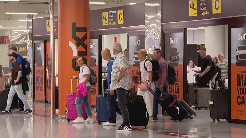 Zona de llegadas de aeropuerto con viajeros y equipaje. Paneles publicitarios de SIXT y señalización multilingüe "Arribades / Arrivals / Ankunft / Llegadas".