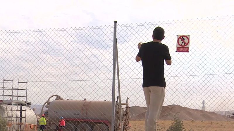 Hombre observa a través de valla con señal de prohibición "PROHIBIDO EL PASO A TODA PERSONA AJENA A LA OBRA". Detrás, sitio de obra con camión cisterna y trabajadores.