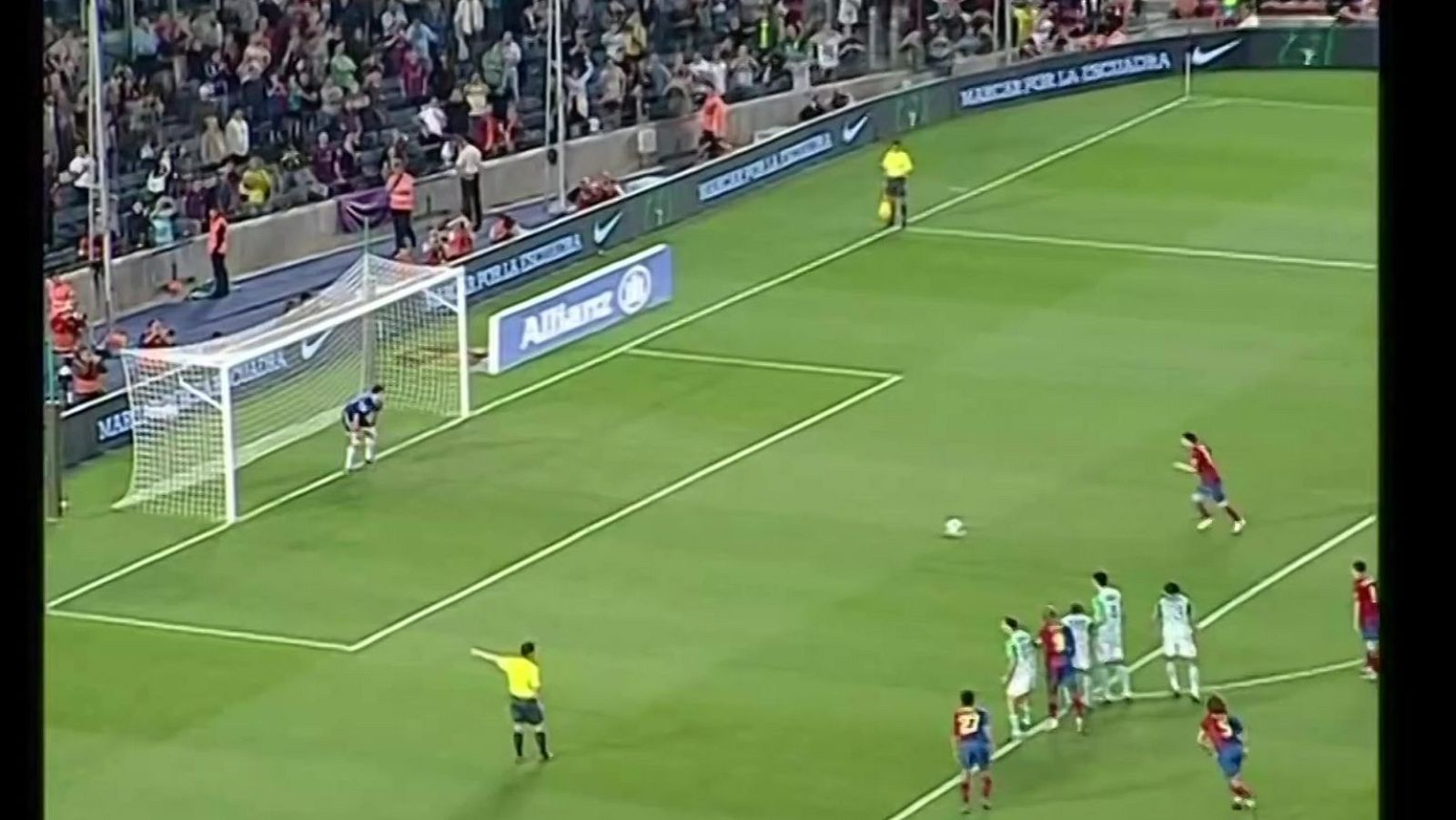 Vista aérea de un tiro libre en un partido de fútbol. Jugador con dorsal 27 del equipo rojiazul frente a barrera blanca y verde. Árbitro amarillo señalando la jugada.