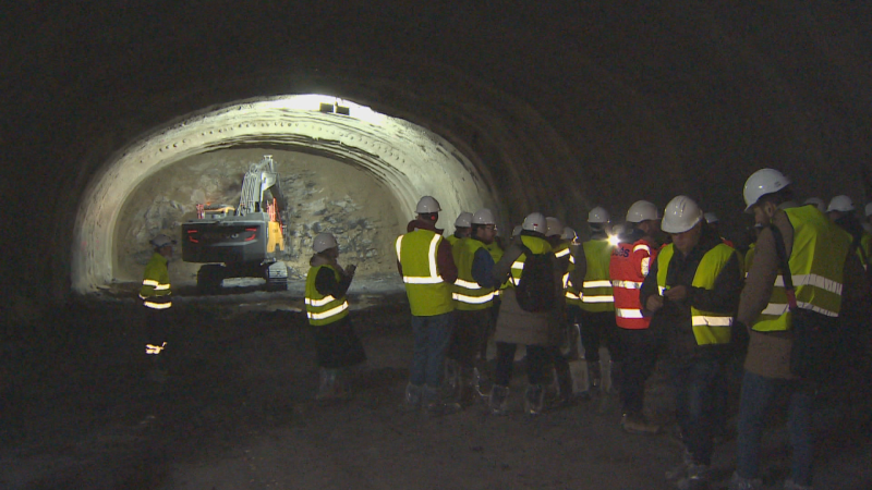 Personas con equipo de seguridad observan un túnel en construcción con maquinaria al fondo. Iluminación tenue y haces de luz de linternas frontales.