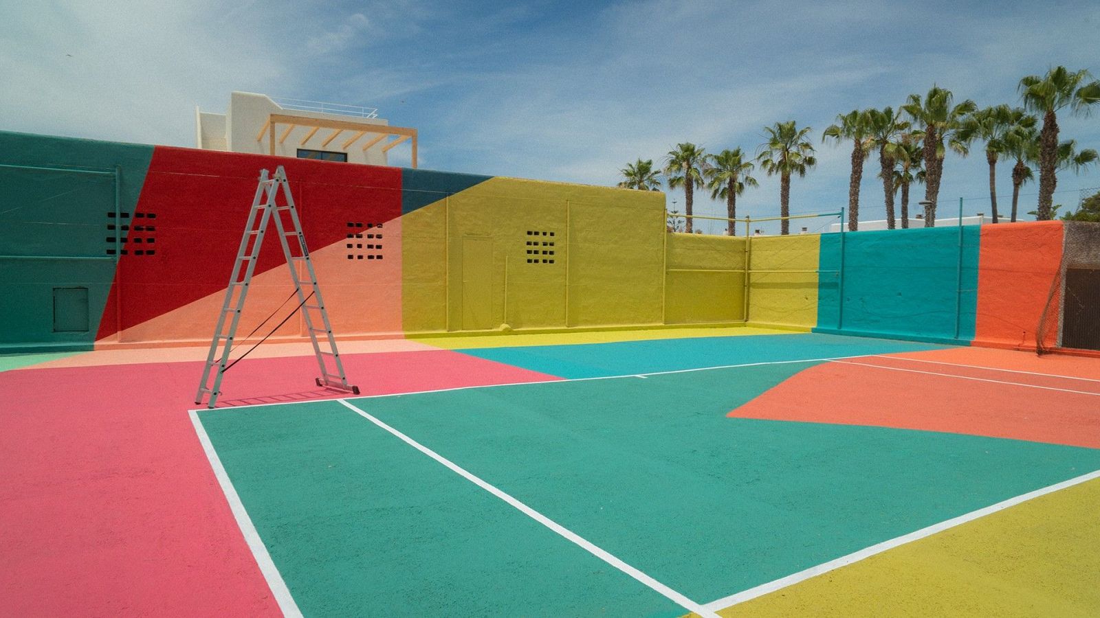Cancha deportiva exterior con diseño geométrico y colores vivos. Escalera de tijera apoyada. Cielo azul y palmeras al fondo.