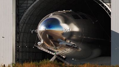 Gran aeronave plateada y reflectante, con h�lices y estructura de soporte, parcialmente dentro de un hangar. Dos personas peque�as visibles en el exterior.