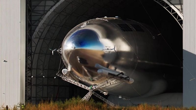 Gran aeronave plateada y reflectante, con hélices y estructura de soporte, parcialmente dentro de un hangar. Dos personas pequeñas visibles en el exterior.