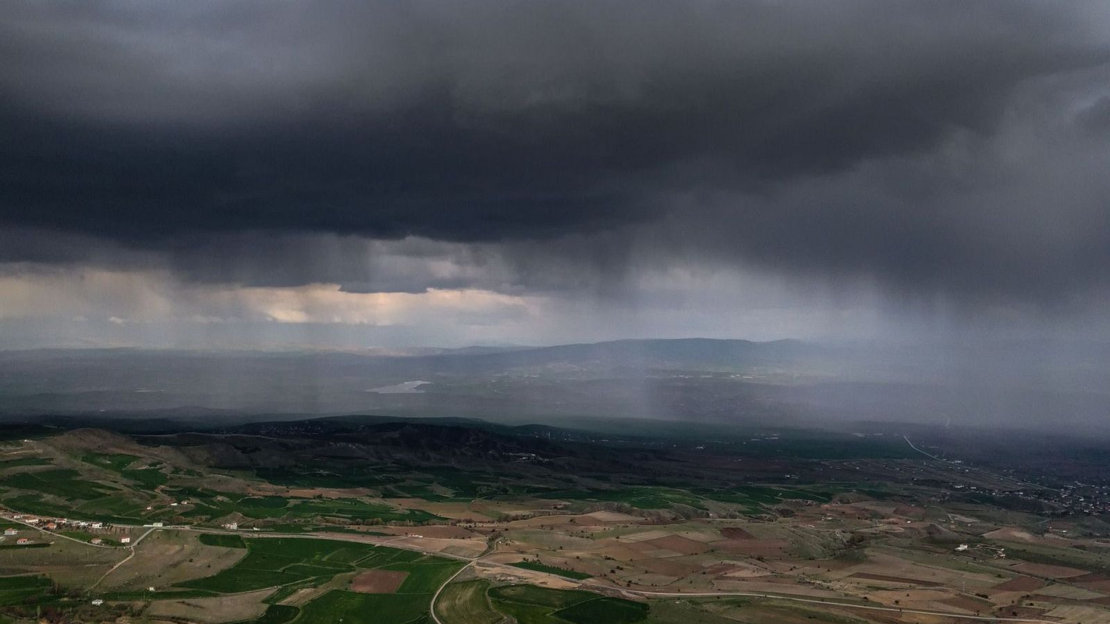 Paisaje rural bajo un cielo tormentoso con lluvia, mostrando campos, colinas y edificaciones.