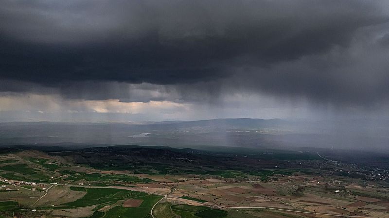 Paisaje rural bajo un cielo tormentoso con lluvia, mostrando campos, colinas y edificaciones.
