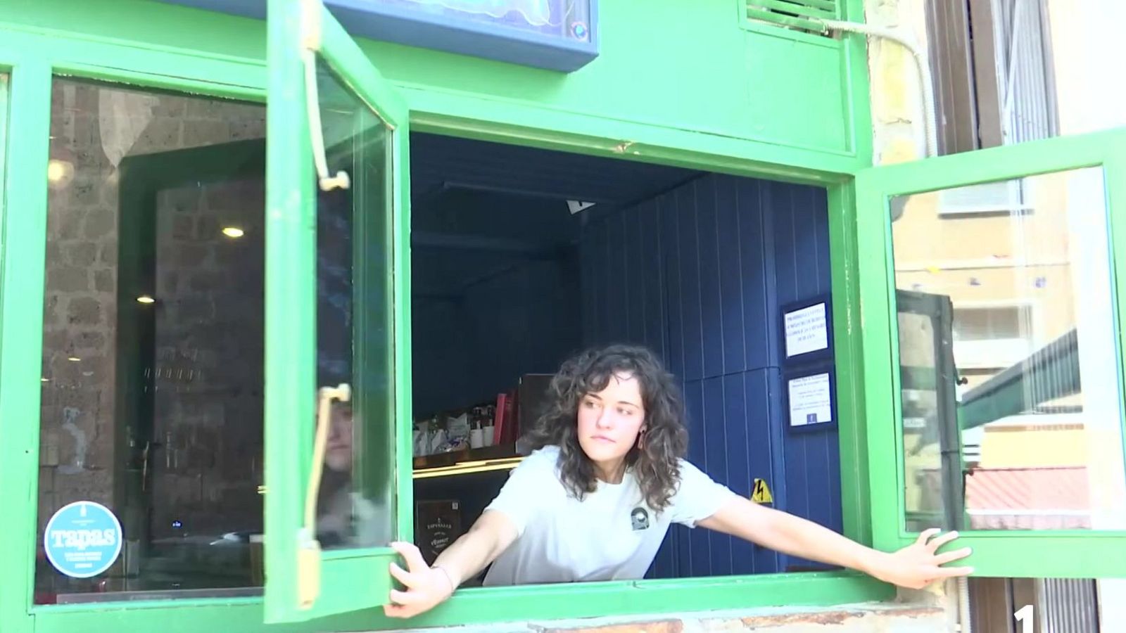 Joven asomada por ventana verde brillante de establecimiento, viste camiseta blanca, mira hacia afuera. "Tapas" visible en cristal.