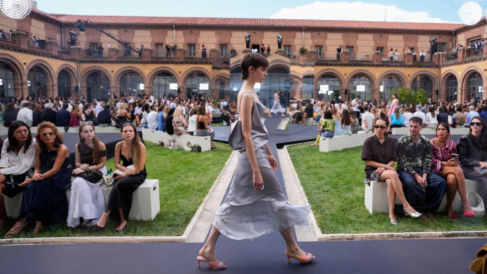 Desfile de moda al aire libre en Madrid. Modelo con vestido plateado satinado camina por pasarela sobre césped. Público observa rodeado de arquitectura histórica.