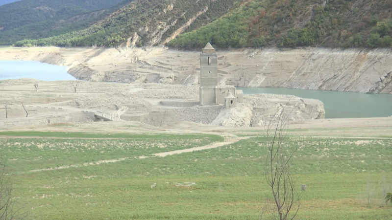 Torre de iglesia de piedra expuesta en un embalse casi seco, rodeada de tierra agrietada y montañas al fondo.