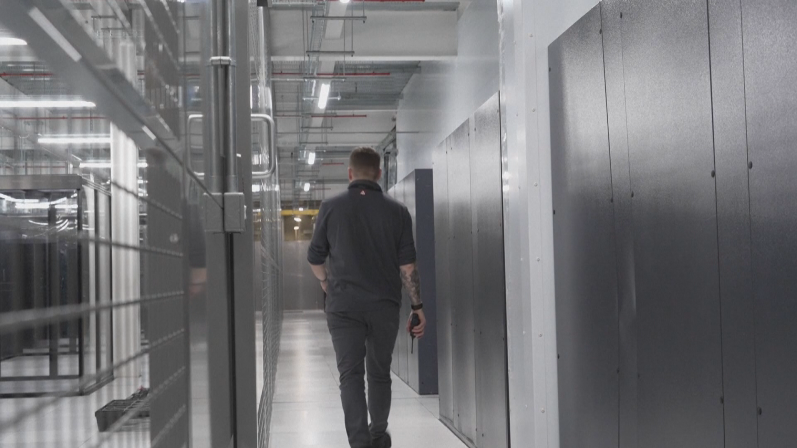 Hombre con tatuajes y walkie-talkie avanza por pasillo de centro de datos entre racks de servidores, bajo iluminación uniforme.