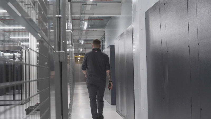 Hombre con tatuajes y walkie-talkie avanza por pasillo de centro de datos entre racks de servidores, bajo iluminación uniforme.
