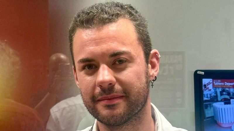 Hombre joven con pendientes en la oreja izquierda, camisa blanca. Fondo dividido: pantalla digital con "Tablero Deport" a la derecha, fondo borroso rojizo/anaranjado a la izquierda.