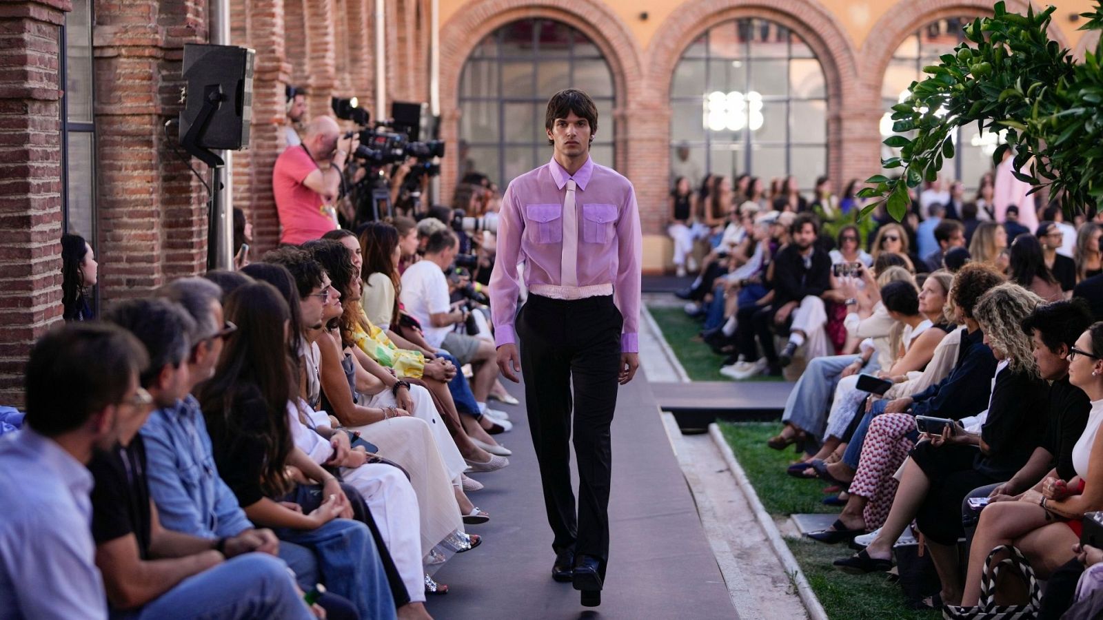 Desfile de moda con modelo masculino en pasarela, arquitectura de ladrillo con arcos y el número "1886" al fondo. Audiencia presente.